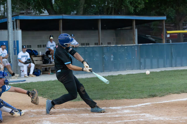 A baseball player in a black uniform swings at a pitch during a game, with spectators visible in the background.