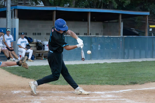 A young baseball player swings at a pitch during a game, with teammates visible in the background.
