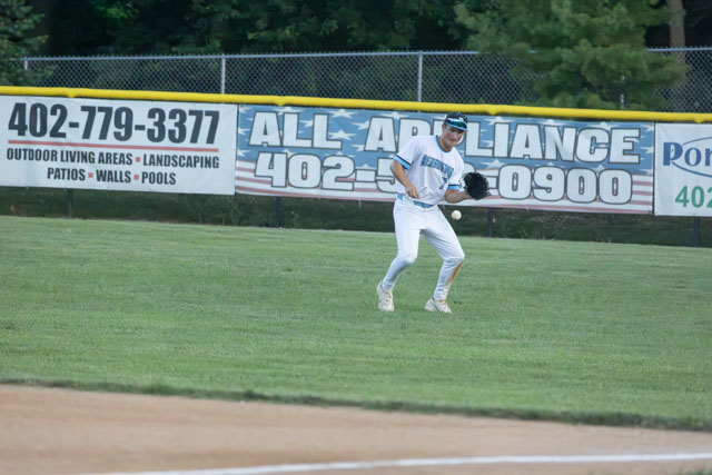 A young baseball player in a white uniform fielding a ball on a grassy baseball diamond, with advertising banners in the background.