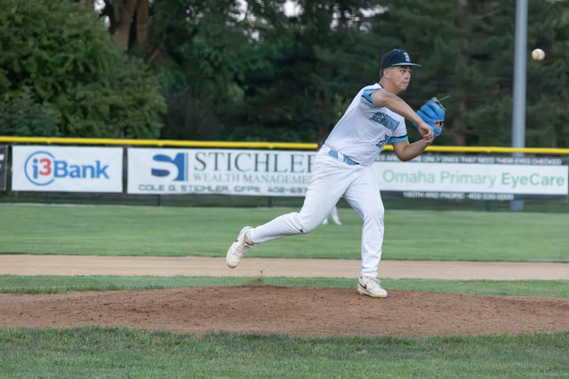 A young baseball pitcher in mid-throw on the pitcher's mound, wearing a blue cap and jersey, with a baseball field and advertisements in the background.