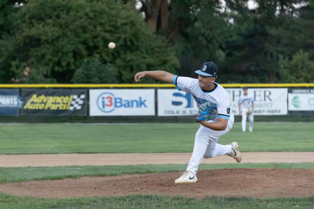 A baseball pitcher in a white uniform throws a pitch on the field, with a blurred ball in motion.