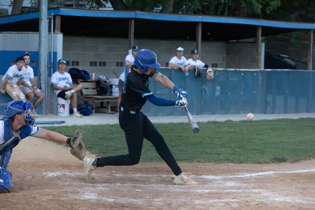 A baseball player swings at a pitch while a catcher prepares to catch the ball behind home plate, with teammates in the dugout in the background.