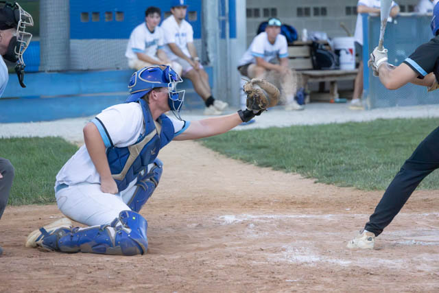A baseball catcher crouches in position, wearing protective gear, while reaching for a pitch. In the background, players are seated on benches, observing the game.