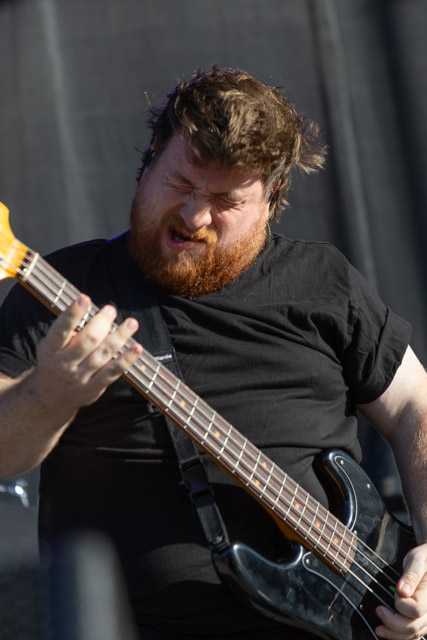 A musician with a thick beard passionately playing an electric bass guitar on stage, with a focused expression and wind-blown hair.