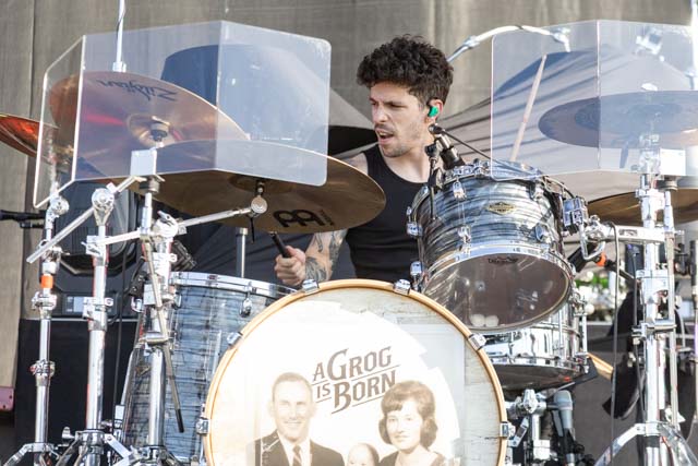 A drummer performing on stage, playing a drum kit with a clear drum shield, wearing an ear monitor and looking focused.