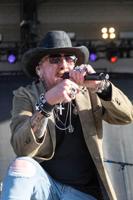 A musician performing on stage, wearing sunglasses and a black cowboy hat. He is holding a microphone and wearing a stylish outfit with multiple bracelets and rings.
