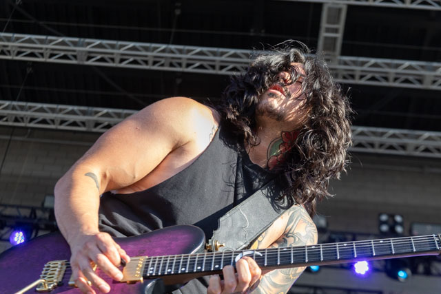 A musician playing an electric guitar on stage, with long, flowing hair and tattoos visible on his arms.