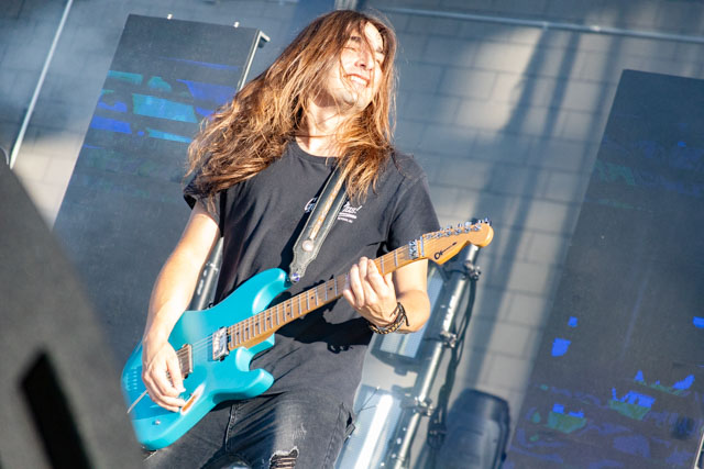 A guitarist with long hair performing on stage, playing a bright blue electric guitar during a live music event.