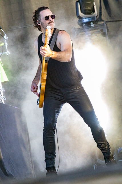 A musician performing on stage, playing an electric guitar, wearing sunglasses and a sleeveless top, with a backdrop of smoke and lights.