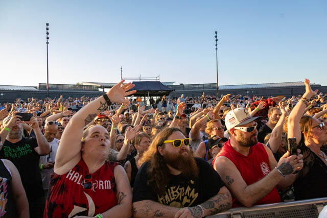 A large crowd of concert attendees with raised hands, enjoying a live performance under a clear sky.
