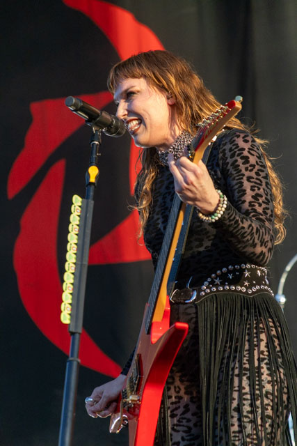 A female guitarist performing on stage, wearing a leopard print outfit and playing a red guitar, with a microphone in front and a colorful backdrop.