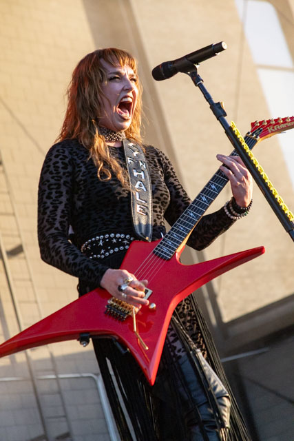 A musician passionately performing on stage, wearing a black leopard-print outfit and playing a red electric guitar.