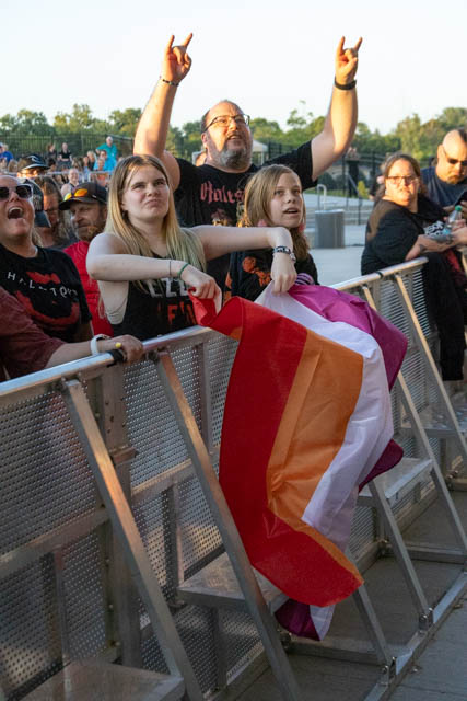 A group of concertgoers at an outdoor event, with one young person holding a pride flag, while others cheer and display excitement.