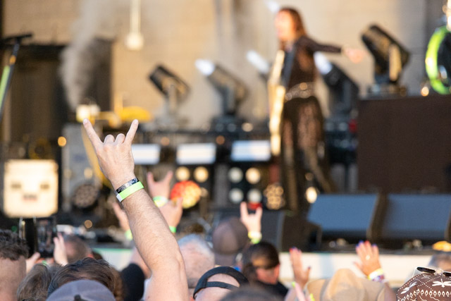 A crowd at a concert with hands raised in a rock gesture, while a performer is on stage in the background.