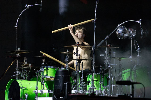 A shirtless drummer energetically playing a bright green drum set on stage, surrounded by instruments and lighting effects.