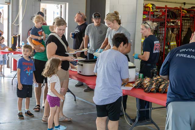A community gathering at a fire station where families are being served food. Adults and children are lined up at a table filled with dishes, with volunteers serving meals. The interior features a casual and welcoming atmosphere.