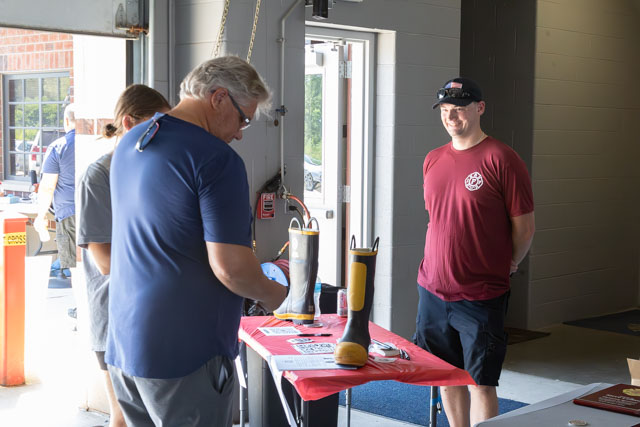 A group of three men interacting inside a building, with one man examining items on a red table. The table displays yellow and black boots, while another man looks on smiling.