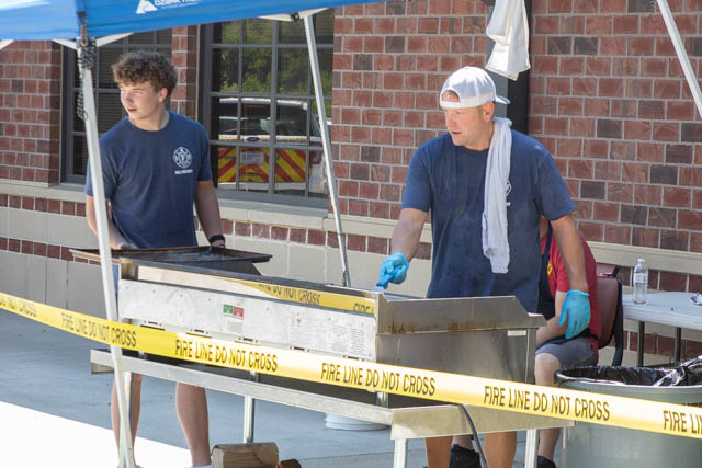 Two young men cooking at a grill station outdoors, with one man flipping food and the other preparing items. They are wearing matching blue shirts and gloves, with a towel around the neck of the man on the right. A yellow caution tape reading 'FIRE LINE DO NOT CROSS' is visible in the foreground.