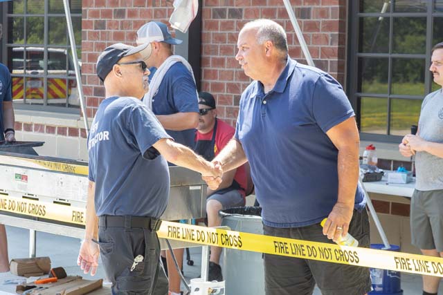 Two men shaking hands at an outdoor event, with a fire line caution tape in the foreground.
