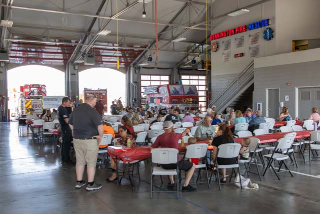 Interior of a fire station during a community gathering, featuring rows of tables with attendees, fire trucks visible in the background, and fire station signage.
