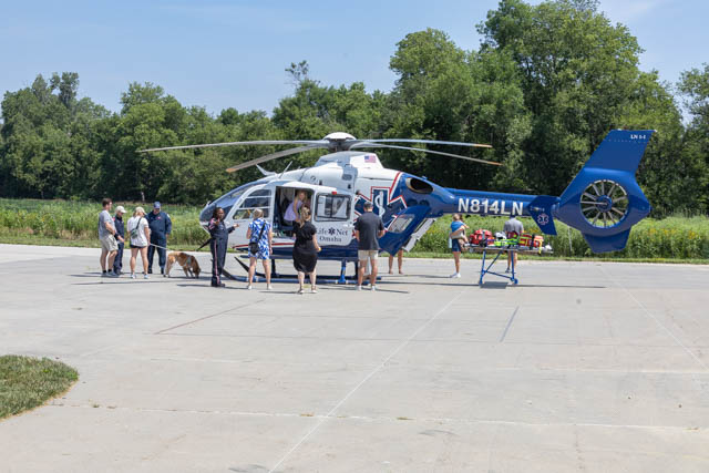 A group of people gathered around a blue and white helicopter on a sunny day, with some individuals interacting with the helicopter crew, while a dog is present among the crowd.