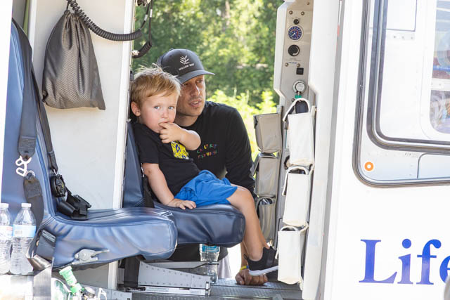 A young boy sitting on a seat inside an emergency vehicle, looking curiously while a man, presumably an adult or parent, sits next to him, both smiling. The scene is set in a bright, outdoor environment.