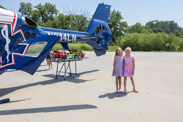 Two young girls standing next to a blue helicopter on a sunny day, with emergency medical equipment in the background.