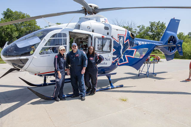 Three medical personnel pose outside a helicopter, wearing flight suits and smiling. The helicopter features medical insignia and is parked on a concrete surface with trees in the background.