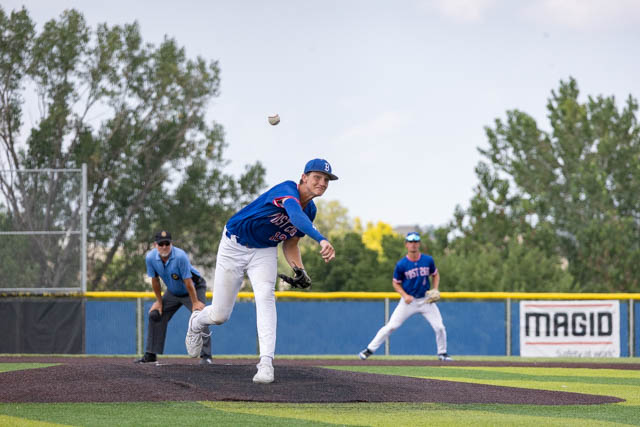 A baseball pitcher in a blue jersey throws a pitch from the mound, while an umpire and a fielder observe in the background.