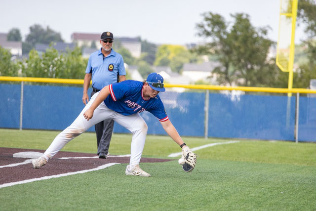 A baseball player in a blue jersey and white pants is fielding a ball on the infield, while an umpire watches from the background.