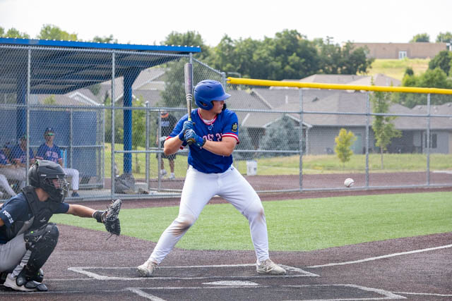 A baseball player in a blue jersey and helmet is preparing to bat as a pitch is thrown, with a catcher in black gear behind him and a baseball field in the background.