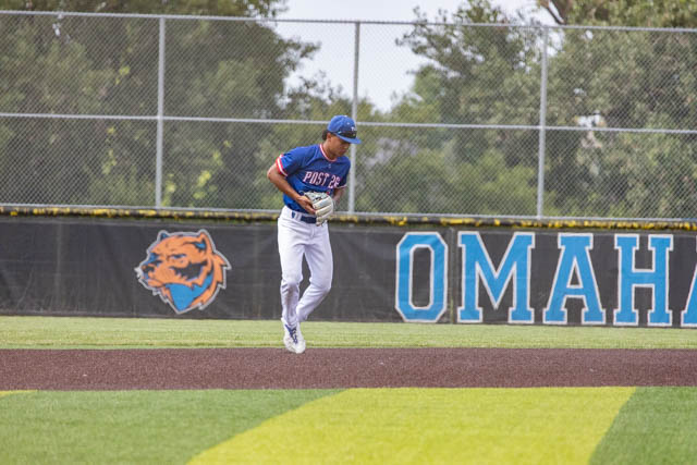A baseball player in a blue cap and jersey running on the field, with a baseball glove in hand, in front of an outfield fence featuring an Omaha team logo.