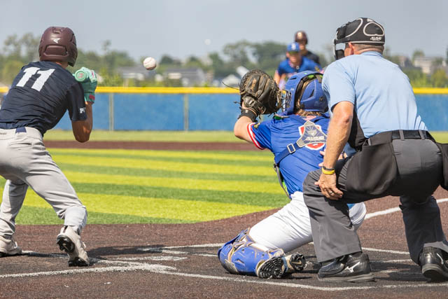 A baseball player in a blue jersey and a catcher crouched behind home plate as a pitch is thrown towards the batter in black and grey uniform. An umpire observes the play from behind.