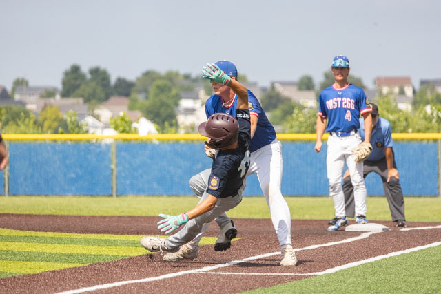 A baseball player sliding into base as a fielder attempts to tag him out during a game. Two other players are visible in the background, and the scene is set on a sunny day.