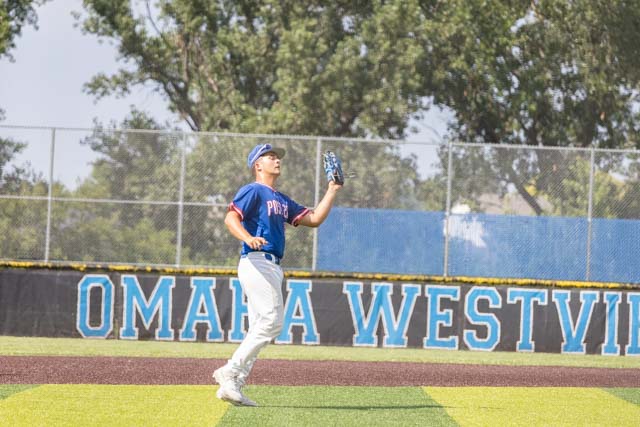 A young baseball player in a blue jersey and white pants is catching a fly ball on a baseball field with a fence in the background that has the words 'OMAHA WESTVILLE' displayed.