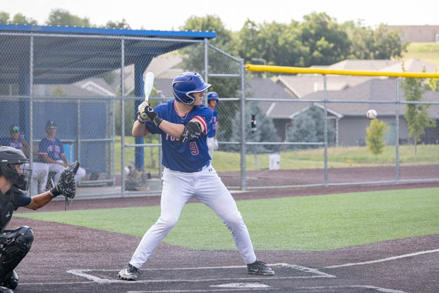 A baseball player in a blue and red uniform stands in a batting stance, preparing to hit a pitch during a game. The scene includes a catcher and players in the background, with a field and houses visible.