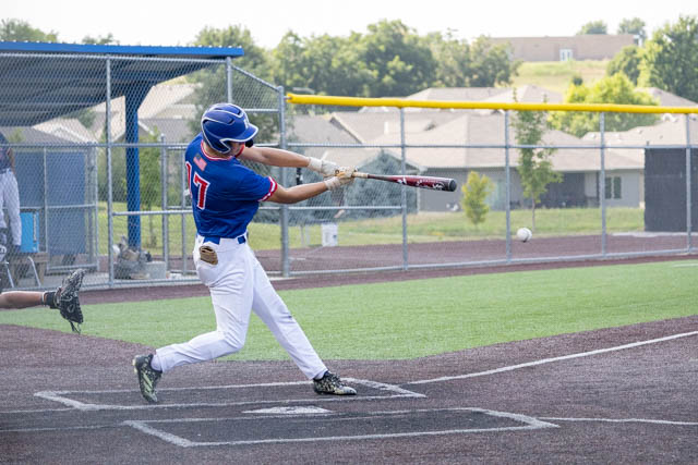 A young baseball player swings a bat at a pitch during a game, wearing a blue jersey with red accents and white pants, on a well-maintained field with a fence and houses in the background.