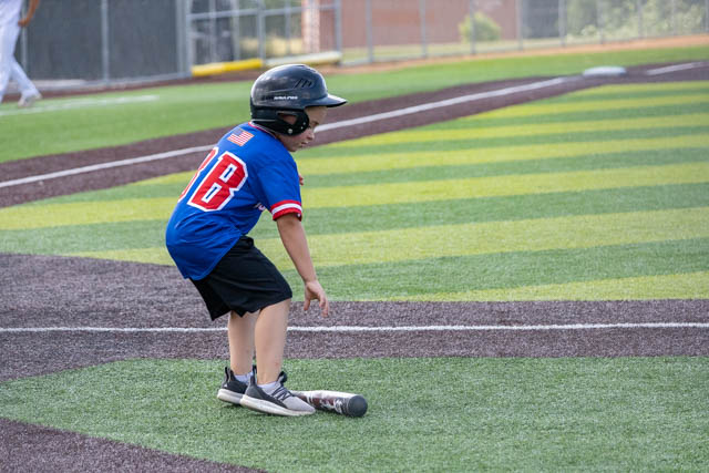 A young baseball player in a blue jersey with the number 18 is crouching near a baseball bat on a green and black turf field.