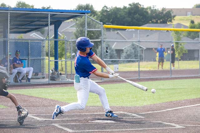 A young baseball player swinging a bat during a game, with players sitting in the dugout in the background.