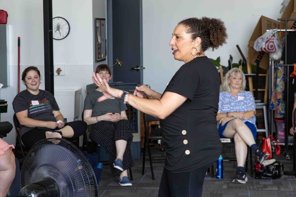 A presenter engages an audience in a workshop, with participants seated and smiling. The room has a casual setting with various objects in the background.