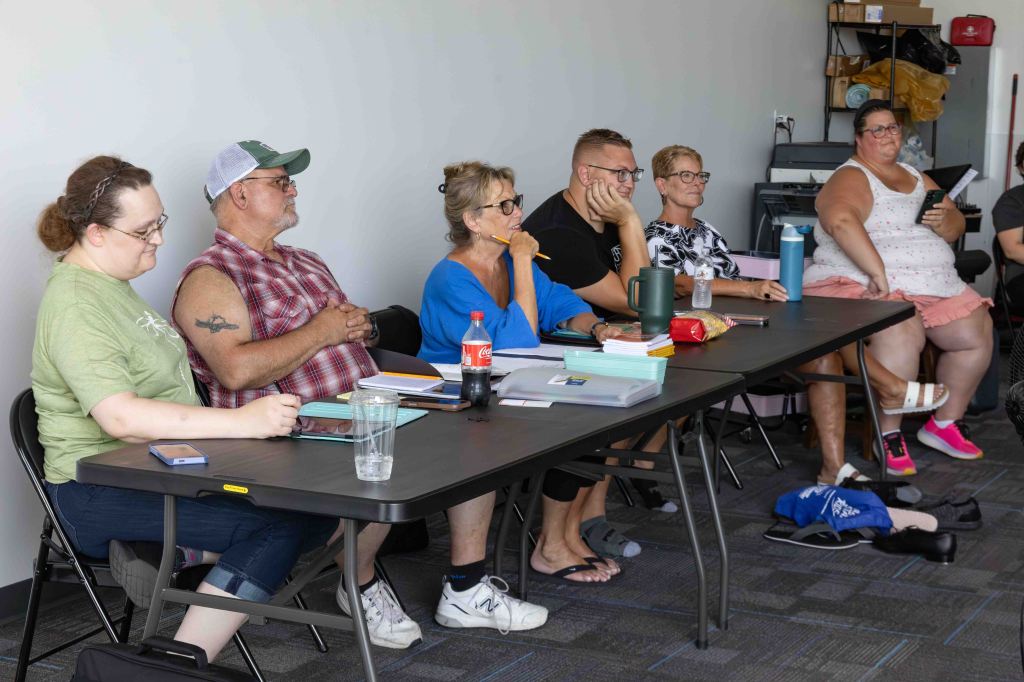 A group of six people sitting at a table in a classroom setting, engaged in a discussion or presentation. The individuals vary in age and appearance, with a mix of casual attire and accessories. Various items such as notebooks and drinks are placed on the table.