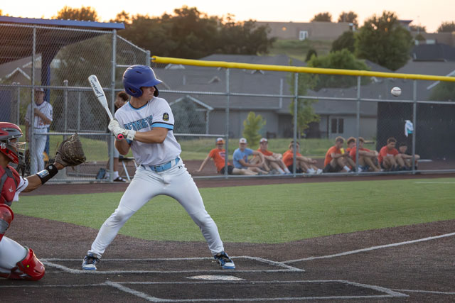 A young baseball player is in a batting stance at home plate, preparing to swing at a pitch during a game. The background features spectators watching the game and a clear sky at sunset.