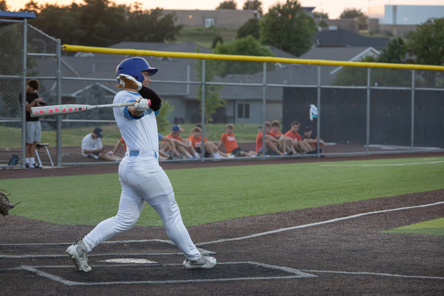 A baseball player in a blue helmet and white uniform swings a bat during a game while a group of spectators sits in the background.