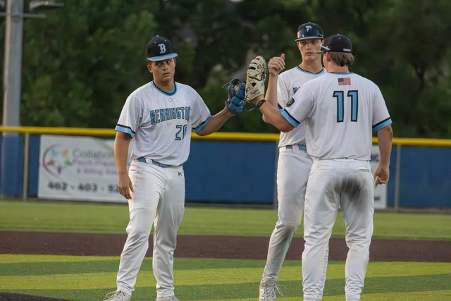 Three baseball players in white uniforms discussing on the field, with one player holding a glove and another wearing a cap.