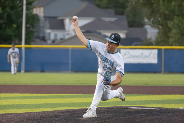 A baseball pitcher in white and blue uniform delivers a pitch on a baseball field, with a blurred player in the background.