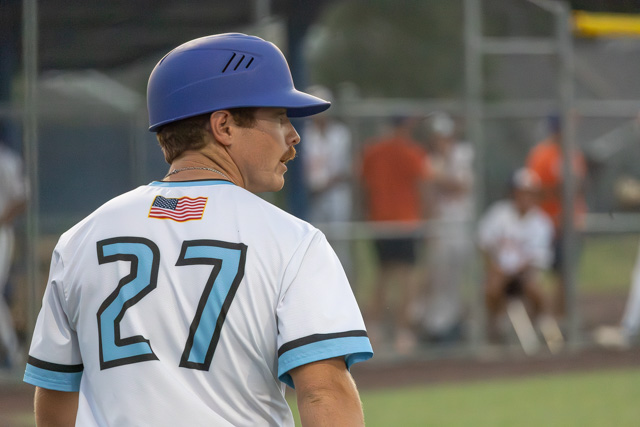 A baseball player wearing a blue helmet and a jersey with the number 27 and an American flag, looking back toward the field during a game.