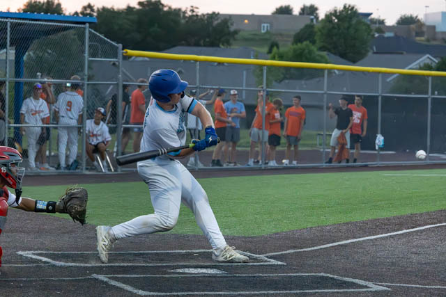 A young baseball player in blue and white swings a bat at a pitch during a game, with a catcher in the foreground preparing to field the ball, and spectators watching from behind.