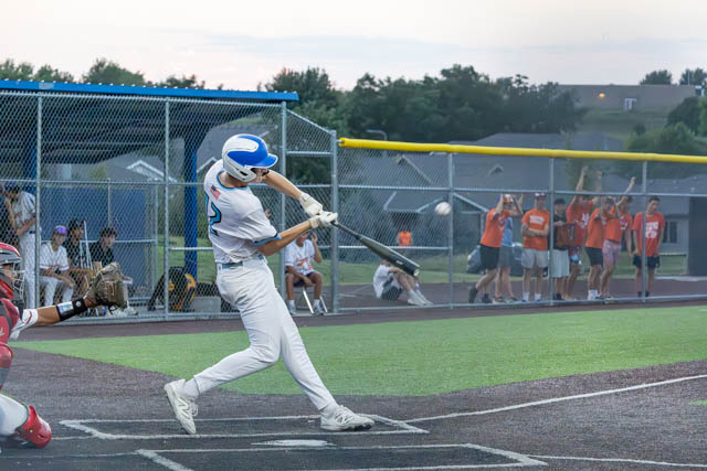 A young baseball player in a white uniform swings a bat during a game, with spectators cheering in the background.