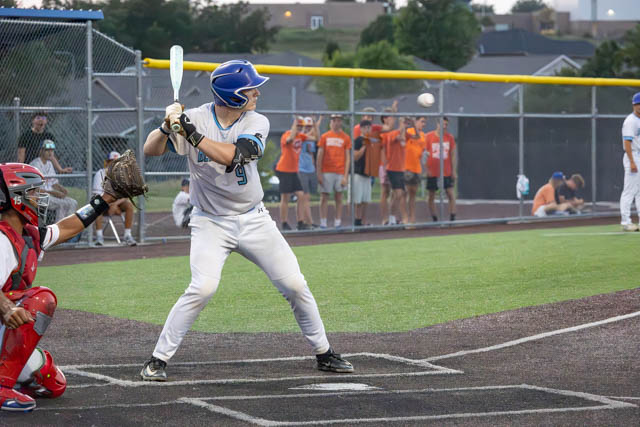 A baseball player in a blue helmet and white uniform stands in the batter's box, preparing to swing as a baseball is pitched towards him. A catcher in red gear is ready to receive the pitch, while a group of fans in the background cheers.