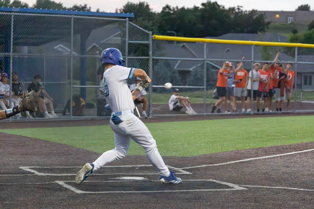 A baseball player in a white uniform swings at a pitch during a game, with fans celebrating in the background.